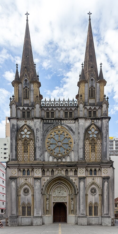 Fachada de uma igreja gótica cinza com duas torres pontiagudas, cada uma com uma cruz no topo. A construção possui três grandes vitrais ornamentados, sendo o central redondo e os laterais em formato de flor, além de uma porta de madeira escura com arco esculpido. O céu azul com nuvens brancas serve de fundo