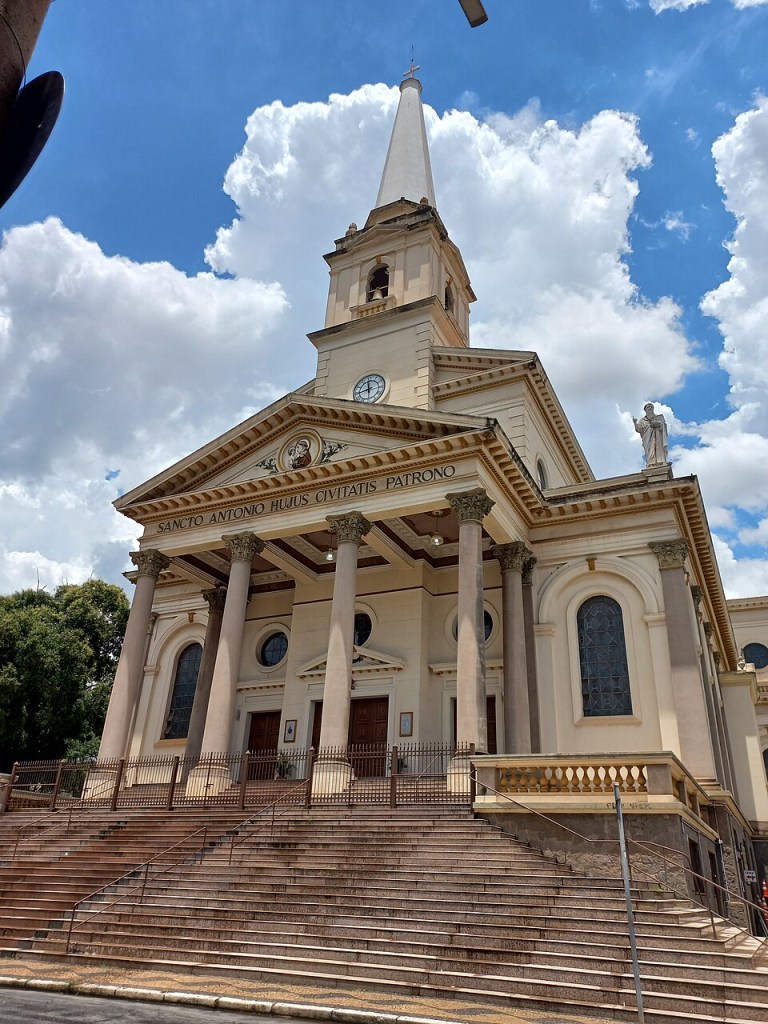 Fachada da Igreja Matriz de Santo Antônio em Americana, com escadaria larga, colunas cor de rosa, torre sineira e céu azul com nuvens brancas