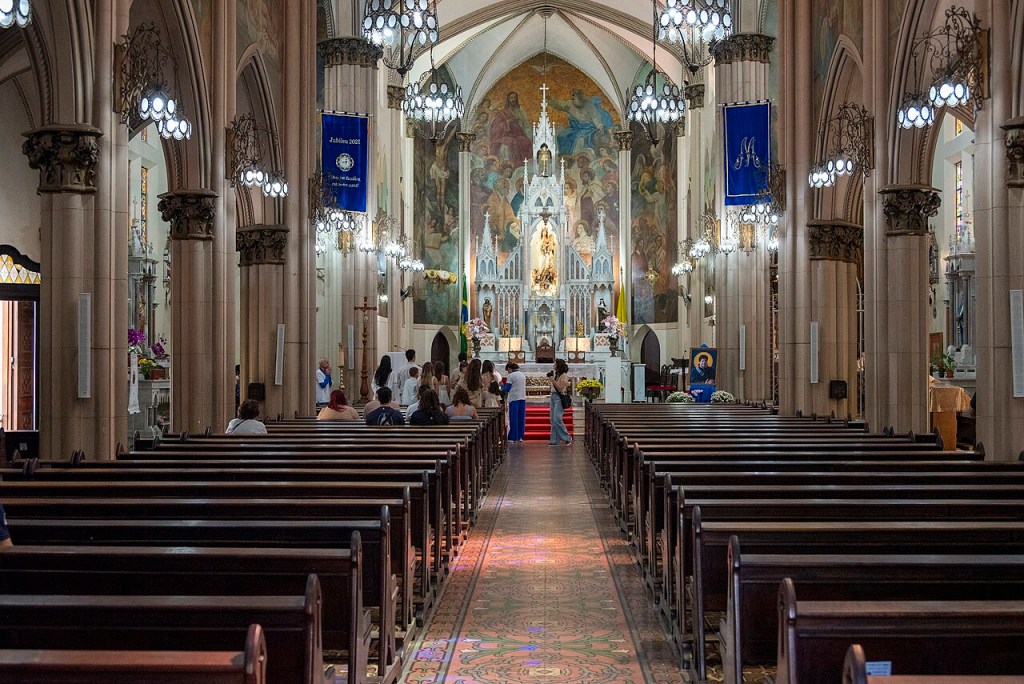 Interior de uma igreja católica com bancos de madeira vazios em primeiro plano, um corredor central com tapete colorido e pessoas próximas ao altar. O altar é branco e ornamentado, com uma imagem de Nossa Senhora e um grande mural pintado ao fundo. Duas bandeiras azuis pendem das colunas laterais, e lustres iluminam o ambiente. Algumas pessoas estão sentadas nos bancos, enquanto outras se aglomeram perto do altar. A arquitetura apresenta arcos e colunas robustas.