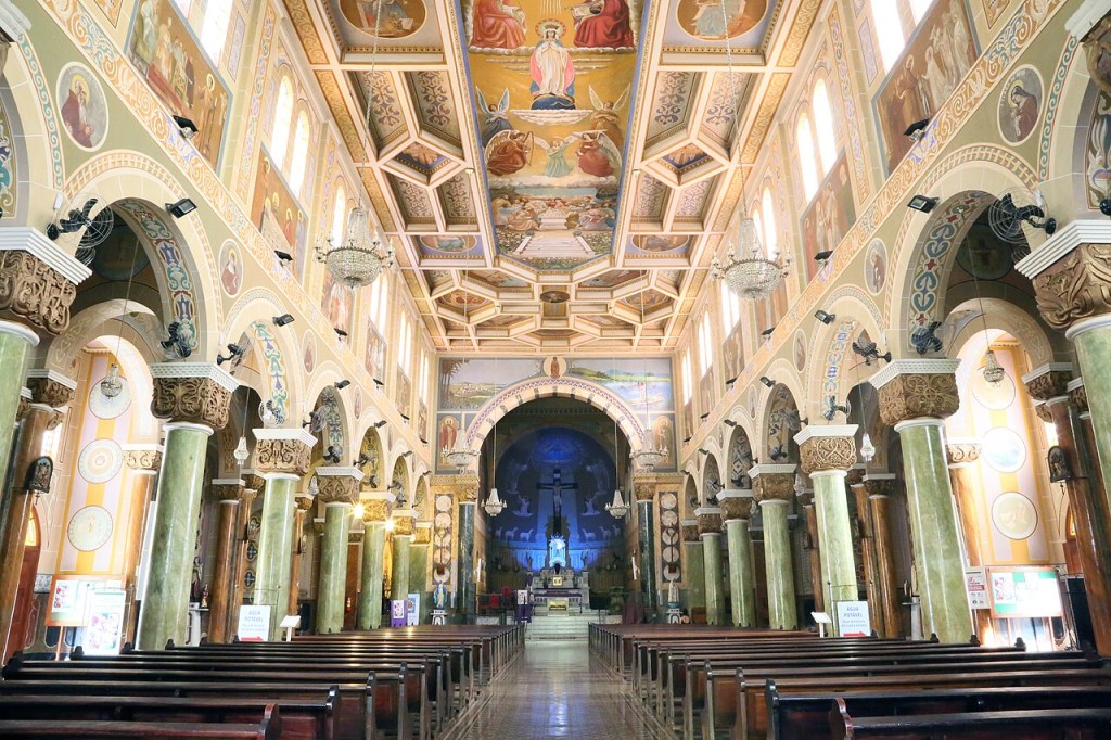 Interior de uma igreja católica com nave central e bancos de madeira. Colunas verdes e arcos decorados sustentam o teto, que exibe afrescos religiosos e lustres. Ao fundo, o altar é iluminado por uma luz azul, com uma cruz e estátuas.