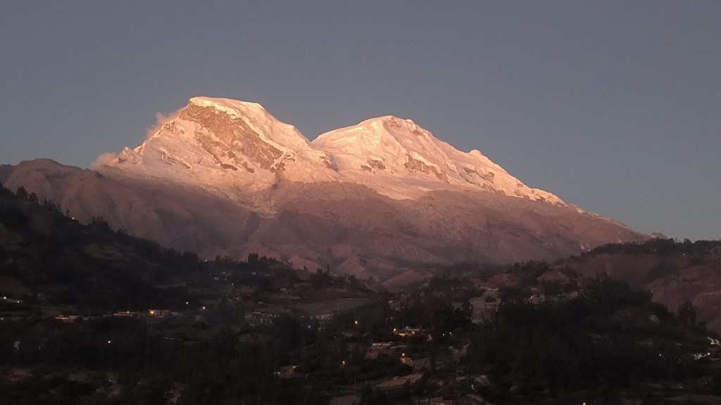 Picos nevados de montanhas iluminados pelo sol da manhã, com encostas rochosas em tons de rosa e roxo. Abaixo, vales escuros com pequenas luzes de casas e vegetação densa, sob um céu azul claro