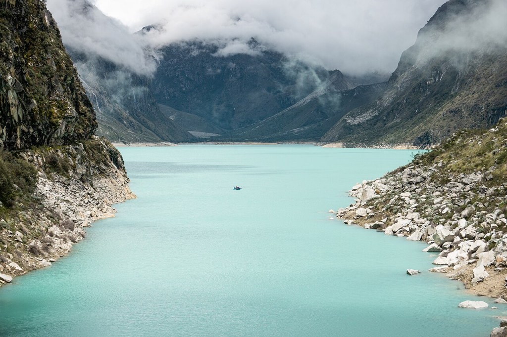 Lago de água azul-turquesa entre montanhas rochosas e íngremes, com nuvens baixas cobrindo os picos. Um pequeno barco azul com duas pessoas navega no centro do lago, sob um céu nublado que cria uma atmosfera serena