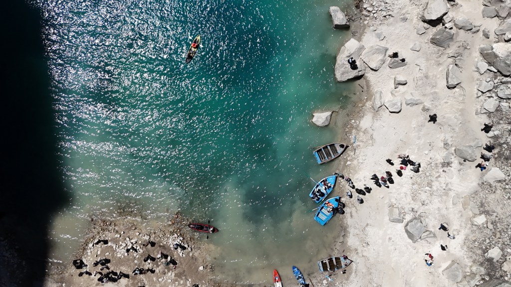 Vista aérea de um lago de águas azul-turquesa e uma margem rochosa. Pessoas em caiaques e barcos azuis navegam no lago, enquanto outras estão na areia e entre as rochas, algumas sentadas, outras em pé. Uma sombra escura cobre parte da água e da margem esquerda