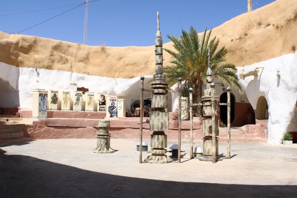 Pátio de um cenário de Star Wars em Tatooine, com construções de barro e brancas, antenas e caixas decoradas com personagens como R2-D2 e Han Solo, sob céu azul