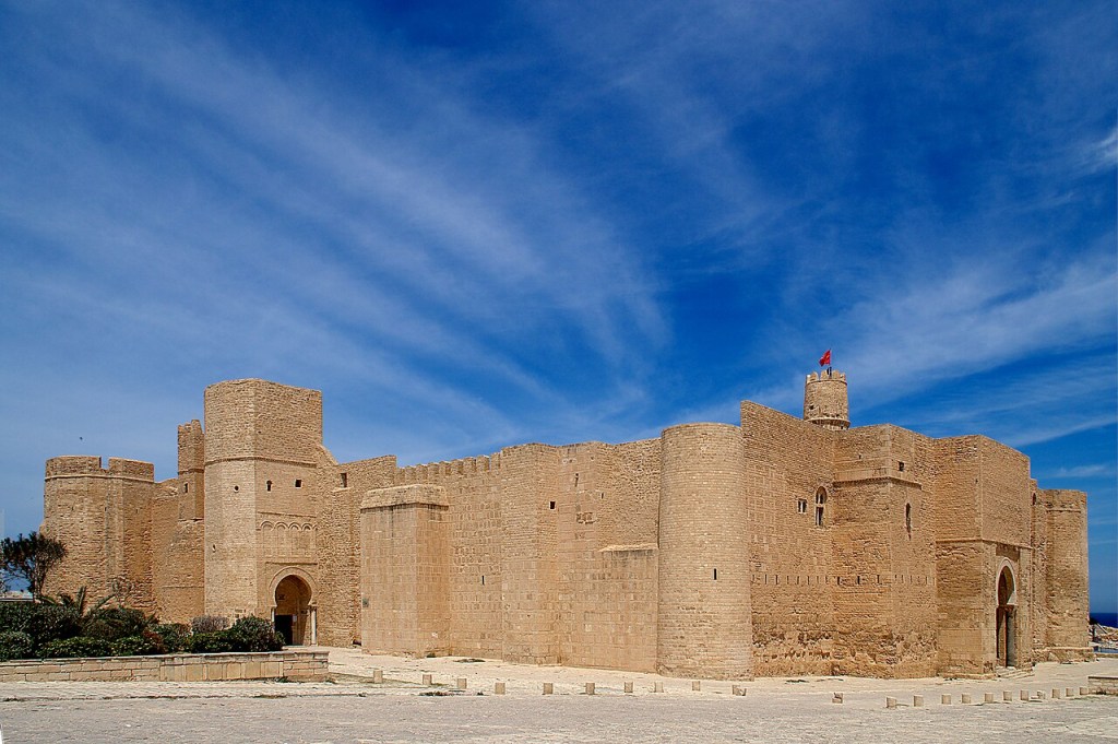 Antiga fortaleza de pedra cor de areia com torres e muralhas, sob um céu azul intenso com nuvens esparsas. Uma bandeira vermelha tremula no topo de uma torre circular.