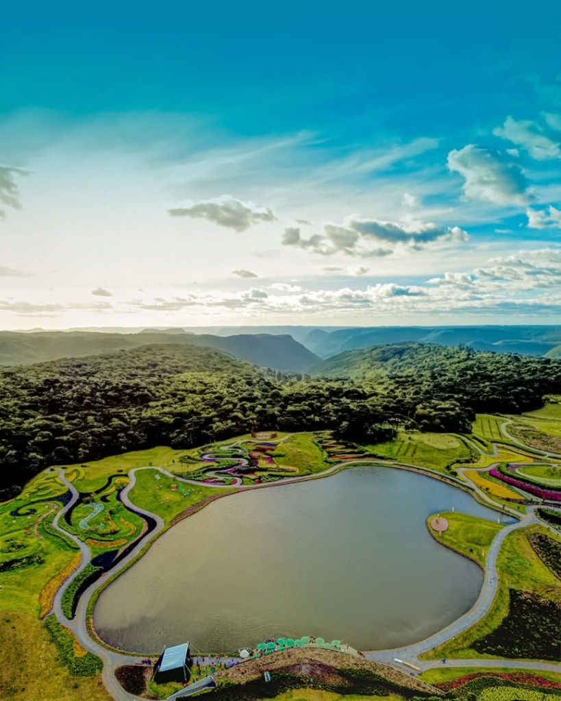 Vista aérea de um lago cercado por jardins coloridos e sinuosos, com trilhas de terra. Ao fundo, uma vasta floresta densa e montanhas sob um céu azul com nuvens esparsas e o sol brilhando no horizonte