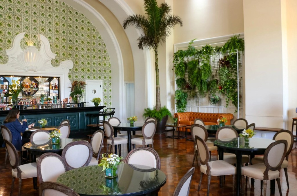 Interior de um bar ou restaurante elegante com piso de madeira, mesas redondas escuras e cadeiras estofadas claras. Ao fundo, um balcão de bar verde-escuro com garrafas e copos, e uma mulher sentada. A parede tem azulejos verdes e brancos, e há um jardim vertical com plantas exuberantes e um sofá de couro marrom