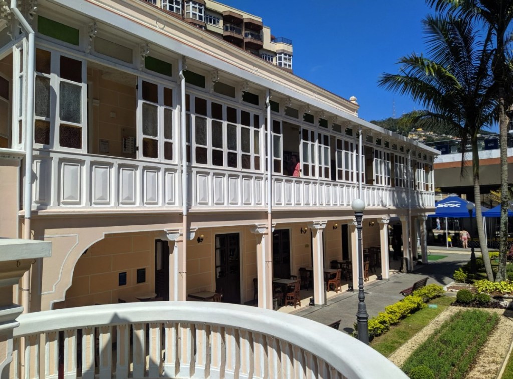 Edifício histórico de dois andares com fachada creme e varanda superior branca, detalhada com balaustrada e janelas de vidro colorido. No térreo, arcos abrigam mesas de café. À direita, palmeiras e um jardim verde sob céu azul. Uma pessoa de camiseta vermelha observa da varanda superior