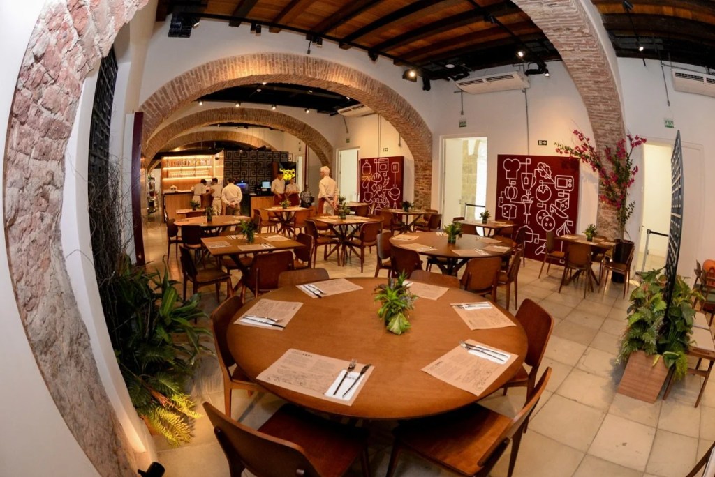 Interior de restaurante com teto de madeira e arcos de tijolos aparentes, mesas redondas e quadradas de madeira escura com cadeiras, e plantas verdes. Ao fundo, funcionários trabalham atrás de um balcão iluminado, e uma parede exibe um painel vermelho com desenhos de utensílios de cozinha