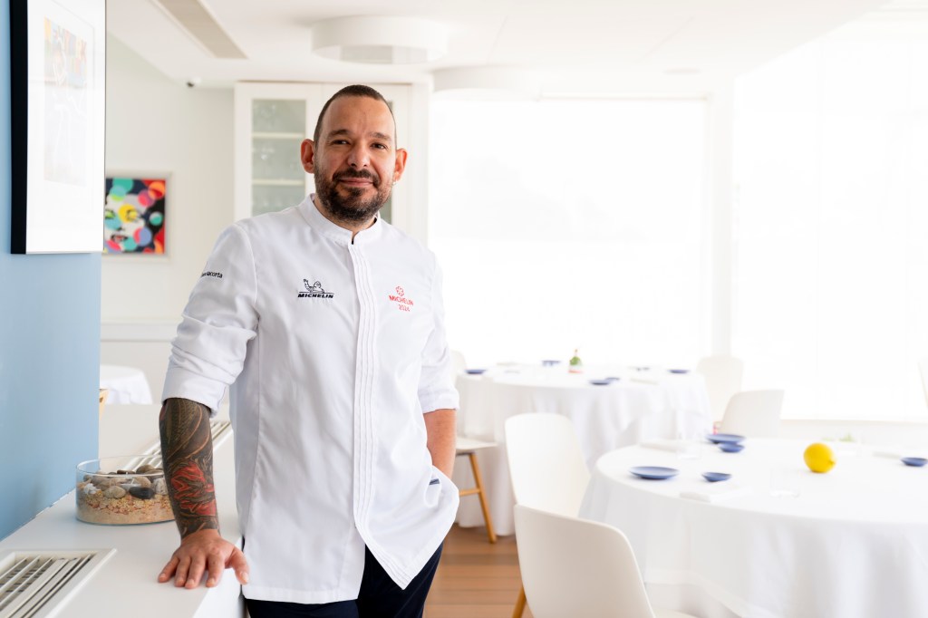 Chef de barba e tatuagem no braço, vestindo dólmã branco com logotipos Michelin, sorri para a câmera em um restaurante iluminado com mesas redondas e toalhas brancas