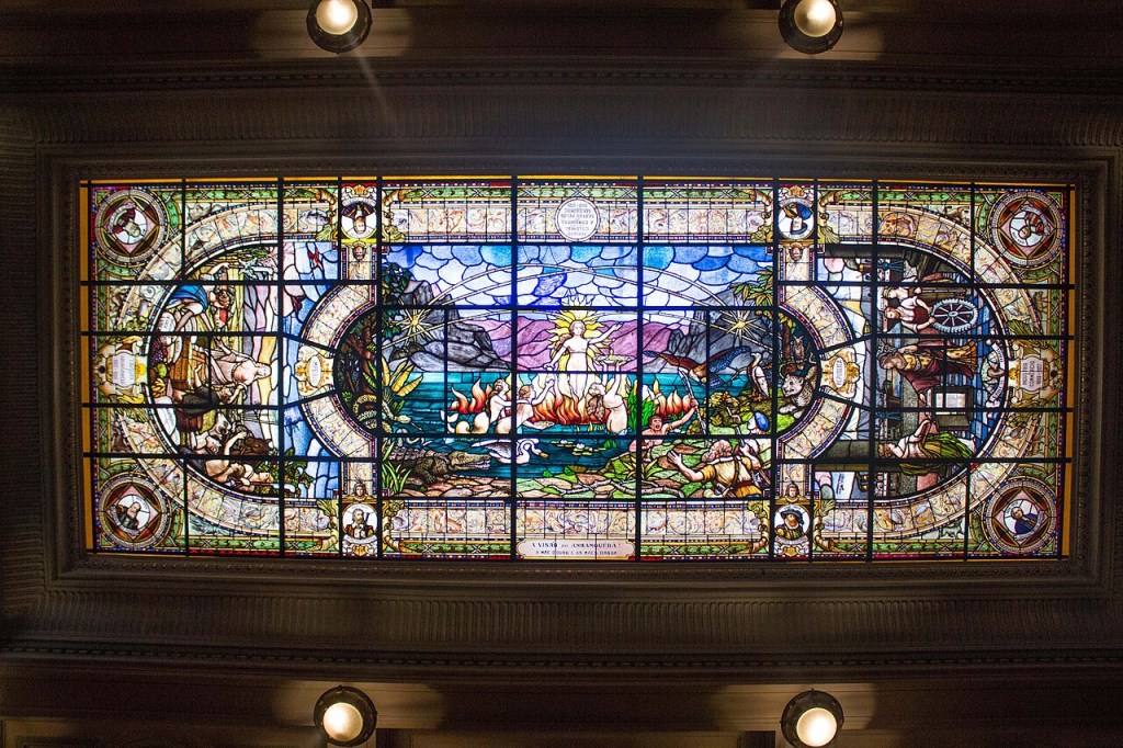 Vitral retangular colorido no teto, com uma cena central oval de uma figura feminina alada sobre um lago com pessoas em chamas e um jacaré. Ao redor, painéis menores retratam figuras históricas e alegorias, como um homem com um livro e outro com uma roda dentada, emoldurados por detalhes ornamentados. A iluminação superior realça as cores vibrantes do vidro.