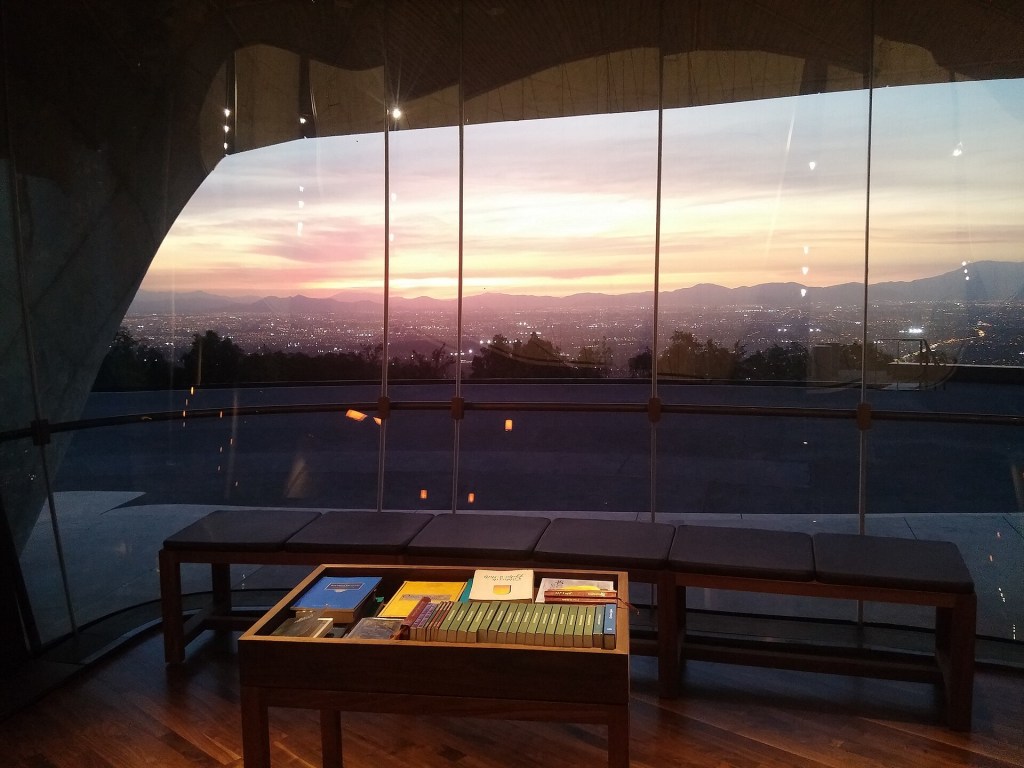 Vista panorâmica de uma cidade ao entardecer, com o céu em tons de rosa e laranja. Em primeiro plano, uma sala com piso de madeira, bancos estofados e uma mesa de centro com livros, tudo através de uma grande janela de vidro que revela a paisagem urbana iluminada e montanhas ao fundo