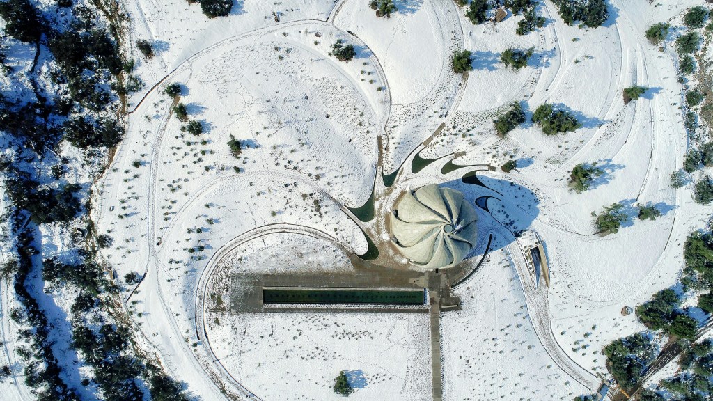 Vista aérea de um templo Baháí em formato de flor, com pétalas cinzas, cercado por caminhos sinuosos e árvores esparsas, tudo coberto por neve branca