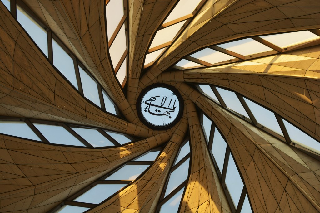 Teto de uma estrutura arquitetônica moderna, com painéis triangulares dourados que se curvam em espiral, formando uma cúpula. No centro, um círculo com caligrafia árabe preta sobre fundo azul claro. Janelas retangulares e trapezoidais entre os painéis revelam o céu azul. A iluminação dourada sugere luz solar.