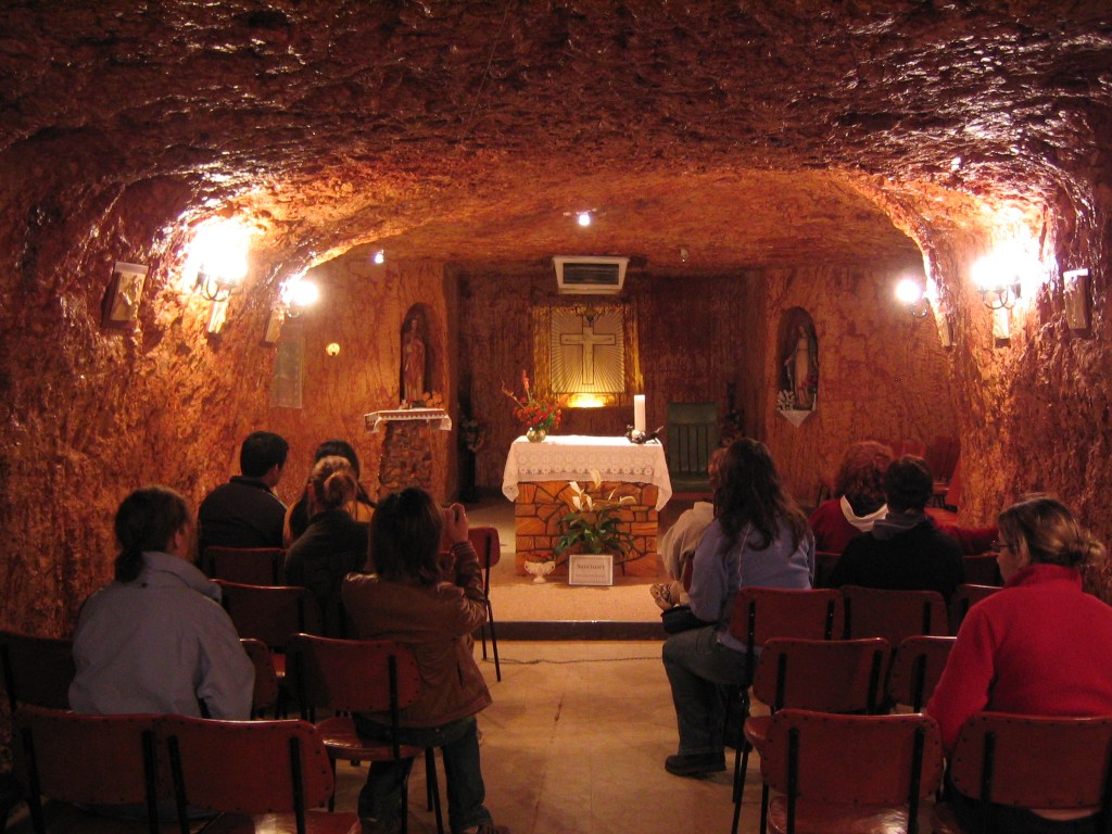 Pessoas sentadas em cadeiras vermelhas dentro de uma igreja subterrânea, com paredes e teto de rocha marrom-alaranjada. Ao fundo, um altar com toalha branca, uma cruz dourada iluminada e uma vela acesa. Há nichos com estátuas de santos e luminárias nas paredes rochosas, criando um ambiente acolhedor e místico