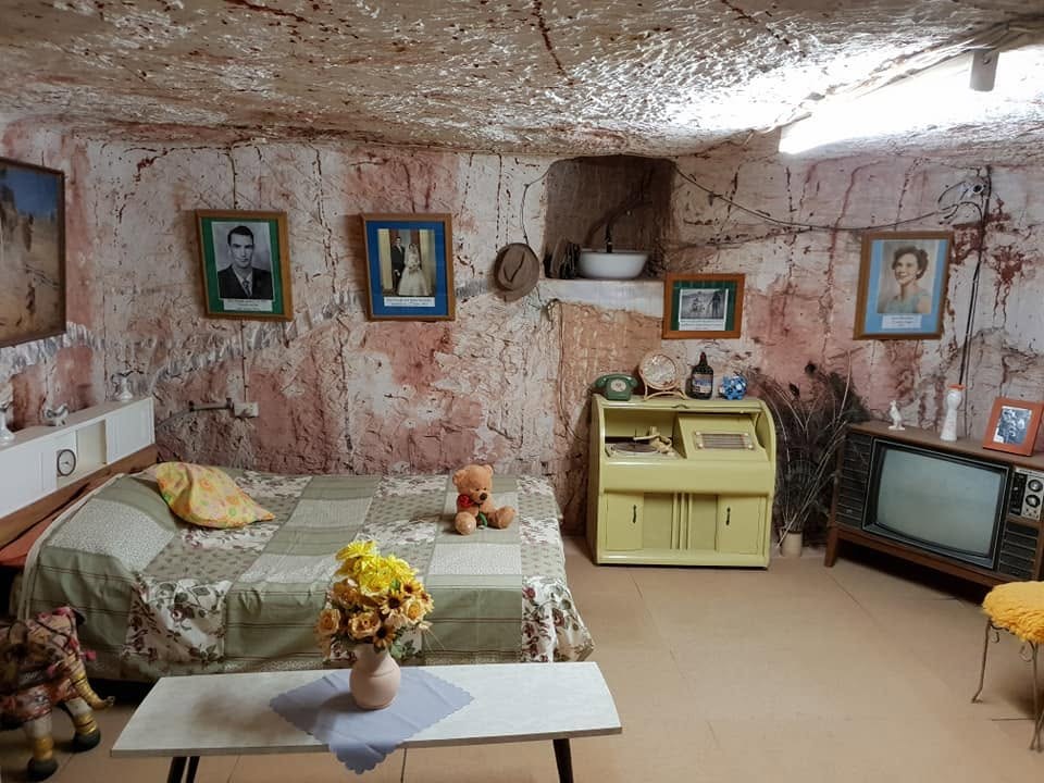 Quarto subterrâneo com paredes e teto de rocha irregular, cama de casal com colcha de retalhos e ursinho de pelúcia, mesa de centro com vaso de flores amarelas, escrivaninha amarela vintage, televisão antiga e fotos de família na parede