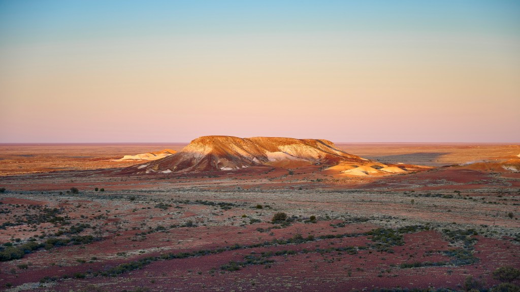 Vasta paisagem desértica com um monte rochoso de topo plano no centro, iluminado por um pôr do sol em tons de rosa e laranja. O solo é avermelhado com vegetação rasteira esparsa, e o céu degrada do azul claro ao rosa pálido no horizonte.