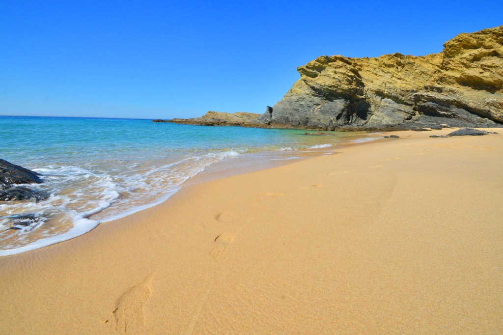 Praia de areia dourada com pegadas, mar azul-turquesa e ondas brancas quebrando na costa. À direita, uma falésia rochosa com tons de cinza e amarelo, sob um céu azul claro