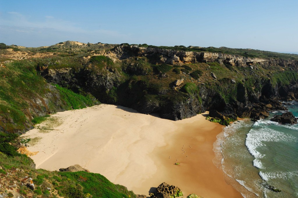 Vista aérea de uma praia isolada com areia clara, cercada por falésias rochosas escuras e vegetação verde. Ondas brancas quebram na orla da praia, e o céu azul claro domina o horizonte