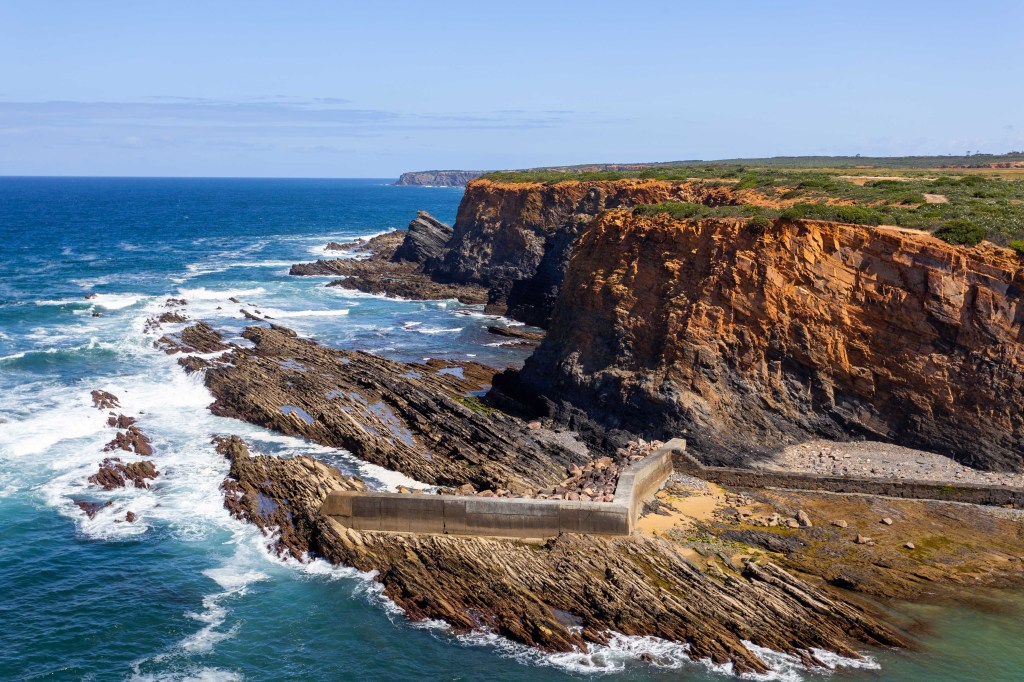 Vista aérea de uma costa rochosa com falésias alaranjadas e vegetação verde no topo, banhada por um mar azul-turquesa com ondas brancas. Uma estrutura de concreto em forma de L se estende da praia para o mar, protegendo uma pequena área de areia e pedras. O céu é azul claro com poucas nuvens