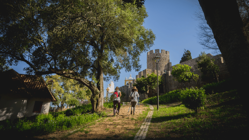 Duas mulheres caminham por uma trilha de terra e tijolos, com bastões de trekking, em direção a um castelo medieval de pedra e uma igreja branca e amarela, sob um céu azul claro. Uma grande árvore frondosa à esquerda e vegetação verde em primeiro plano e ao longo do muro do castelo completam a cena ensolarada