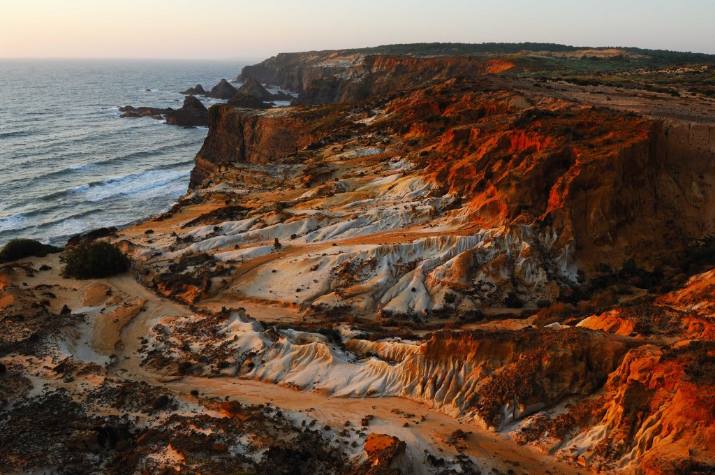 Litoral rochoso com falésias em tons de laranja e branco, erodidas pelo tempo, contrastando com o azul escuro do mar e o céu claro do entardecer