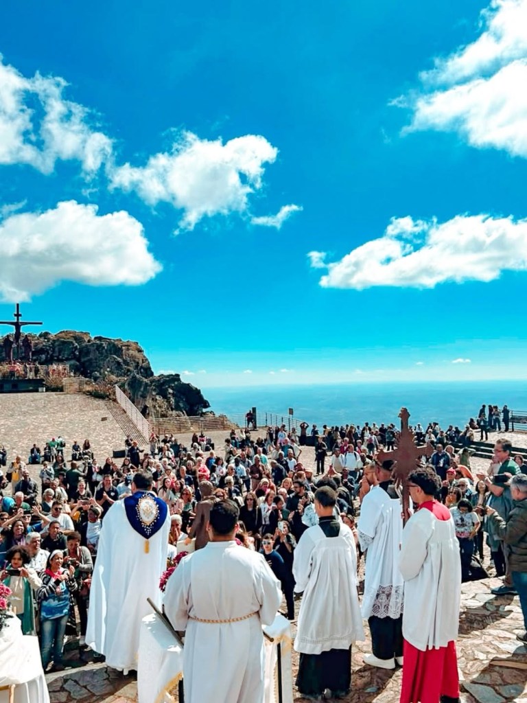Missa campal com sacerdotes de costas para a câmera, celebrando para uma multidão em um mirante rochoso com vista para o oceano azul, sob céu claro com nuvens brancas. Ao fundo, uma grande cruz de madeira no topo da montanha