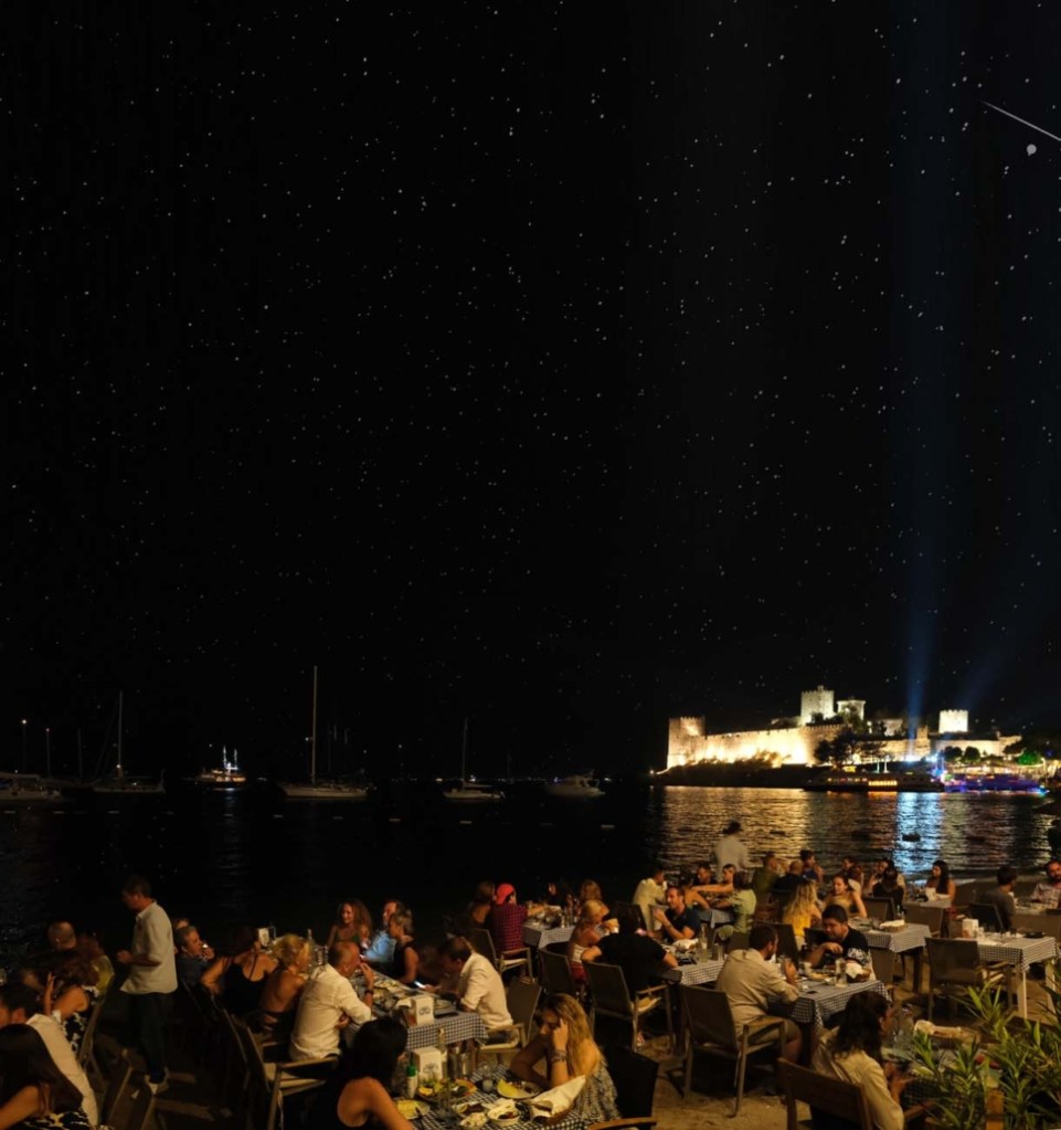 Pessoas jantando em mesas ao ar livre, à beira-mar, sob um céu noturno estrelado. Ao fundo, um castelo iluminado na costa, com barcos ancorados na água escura