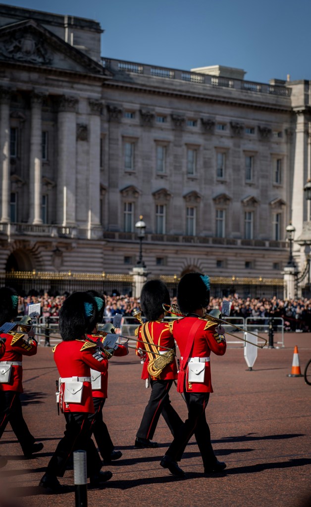 Guardas reais britânicos, de costas, marcham em uniforme vermelho com chapéus de pele preta e instrumentos de sopro, em frente ao Palácio de Buckingham e uma multidão de pessoas