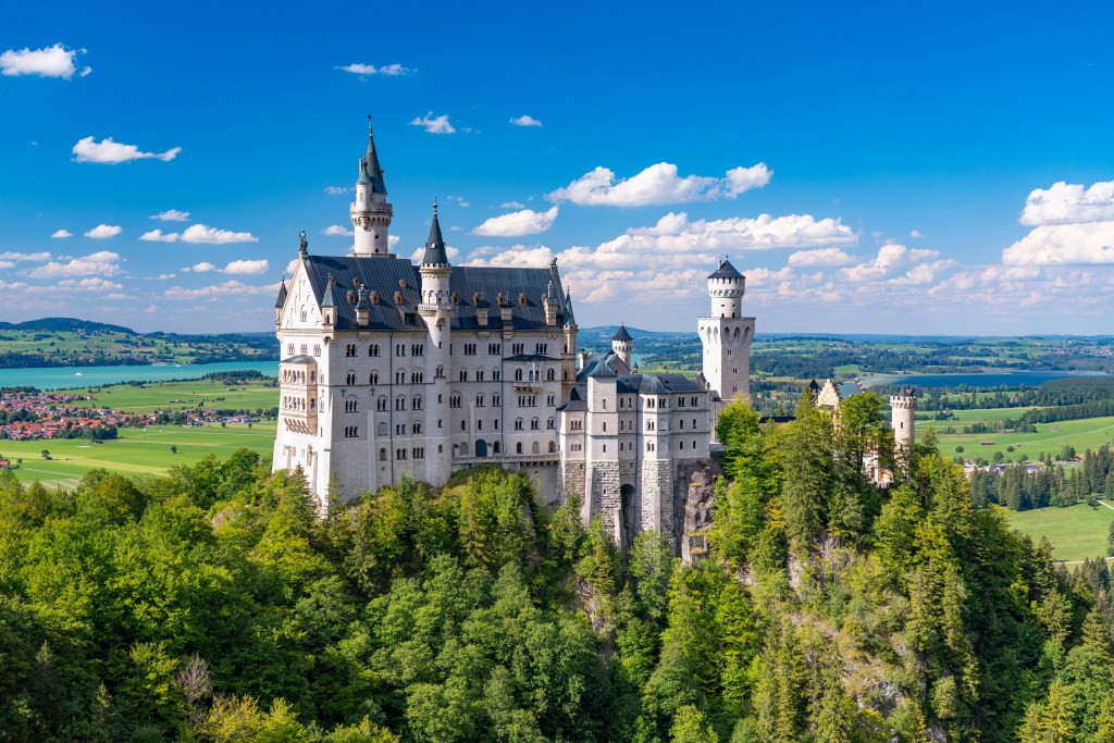 Castelo Neuschwanstein, uma imponente construção branca com telhados escuros e torres pontiagudas, erguido sobre uma colina densamente arborizada. Ao fundo, um lago azul-turquesa e campos verdes se estendem sob um céu azul com nuvens esparsas