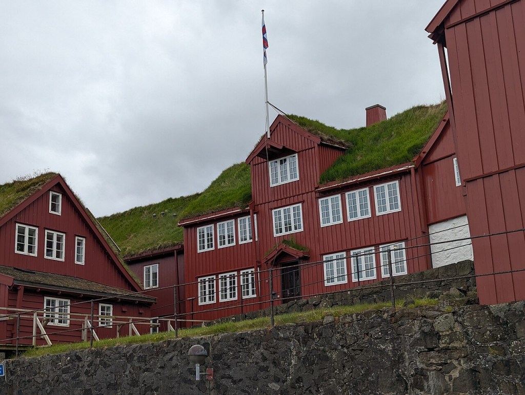 Casas de madeira vermelha com telhados verdes cobertos de grama, algumas com janelas brancas, sob um céu nublado. Uma bandeira tremula no mastro da casa central. Uma parede de pedra escura e um corrimão preto separam as casas do primeiro plano