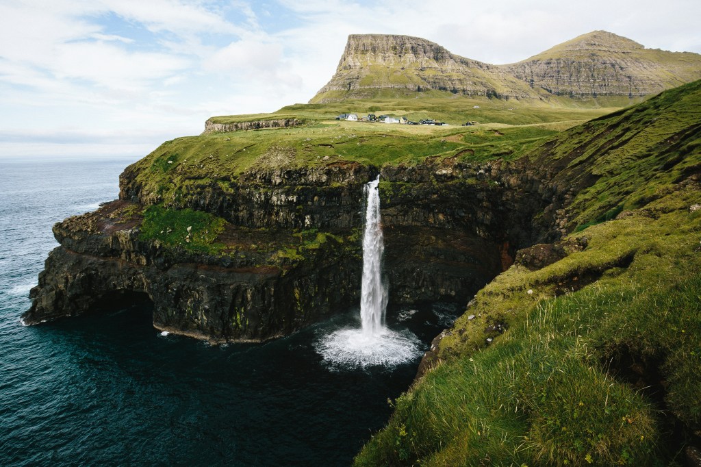 Cachoeira de Múlafossur nas Ilhas Faroé, despencando de um penhasco rochoso coberto de grama verde no oceano azul-escuro. Pequenas casas com telhados escuros ficam no topo do penhasco, sob montanhas rochosas e um céu nublado