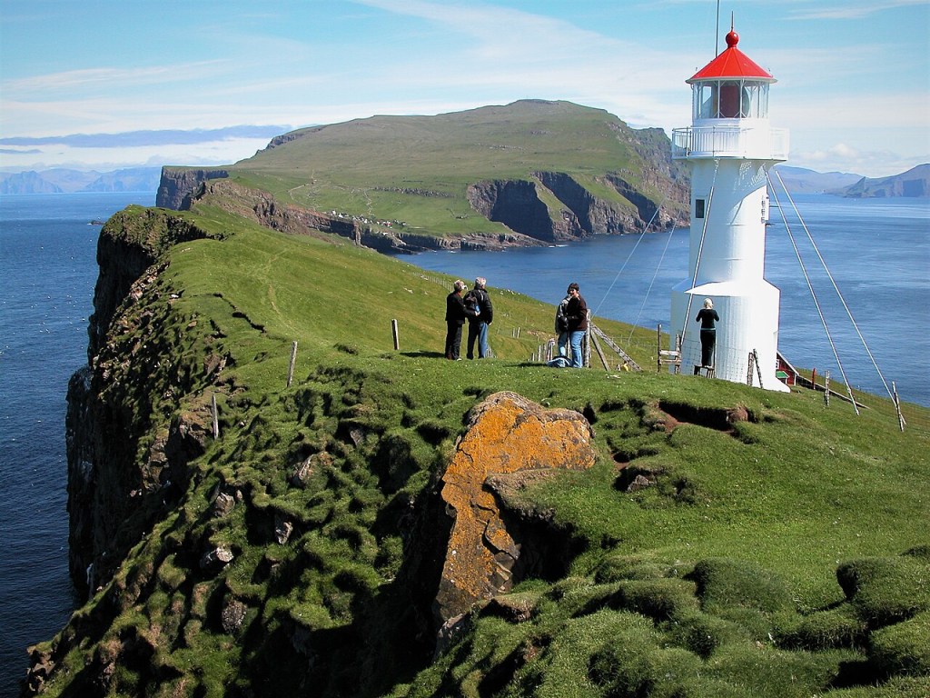 Farol branco com teto vermelho em um penhasco verde, com quatro pessoas observando a paisagem. O mar azul profundo e outras ilhas montanhosas ao fundo completam o cenário ensolarado