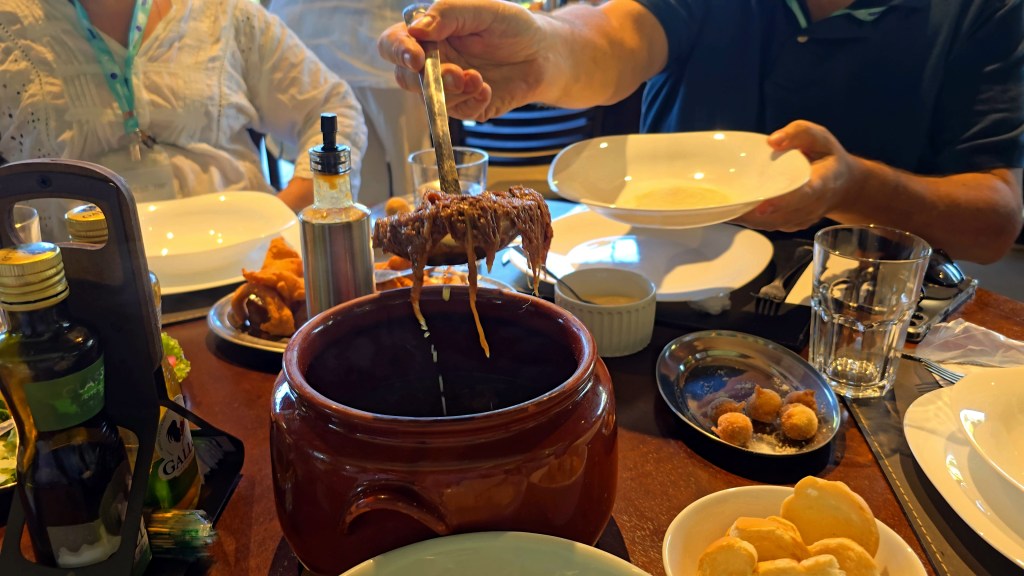 Uma pessoa serve carne desfiada com queijo derretido de uma panela de barro para um prato branco, em uma mesa de madeira com garrafas de azeite, pães e salgados