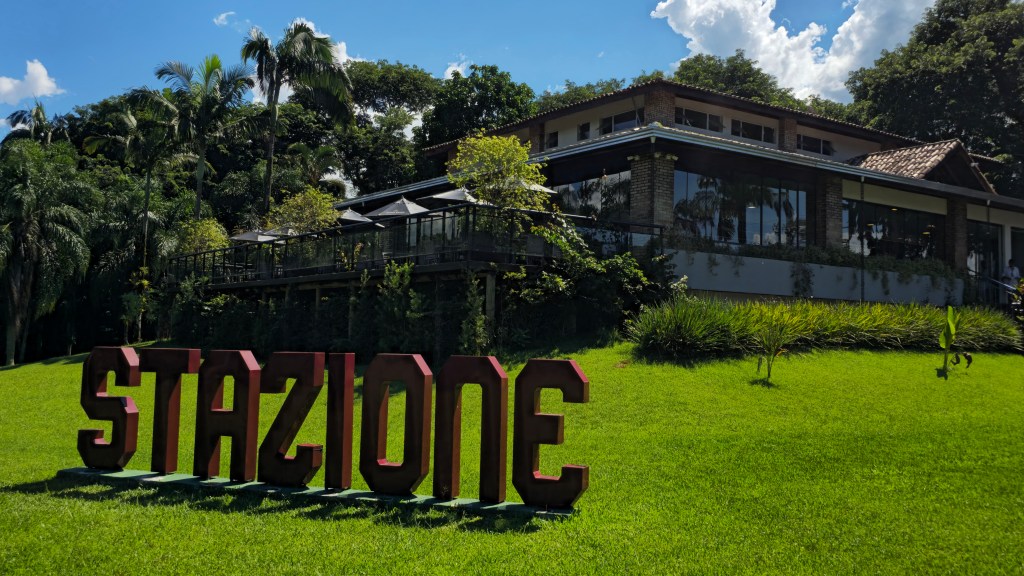 Letreiro vermelho STAZIONE em gramado verde, com um edifício de tijolos e janelas espelhadas ao fundo, cercado por árvores e vegetação exuberante sob céu azul com nuvens