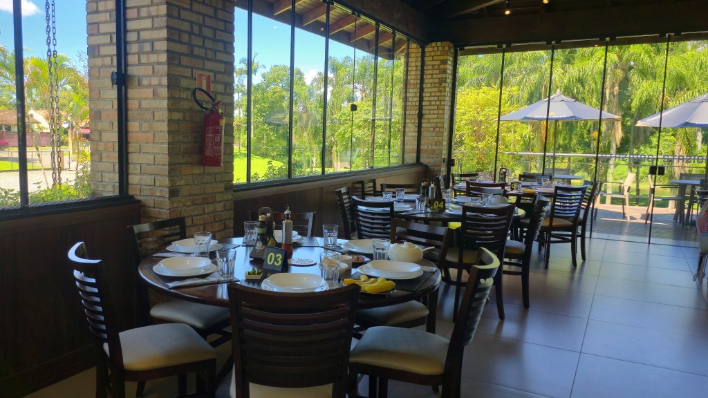 Interior de um restaurante com mesas redondas de madeira escura e cadeiras estofadas, prontas para refeição. Grandes janelas de vidro revelam um exuberante cenário verde com árvores e um lago ao fundo, sob um céu azul. Uma coluna de tijolos aparentes e um extintor vermelho são visíveis à esquerda