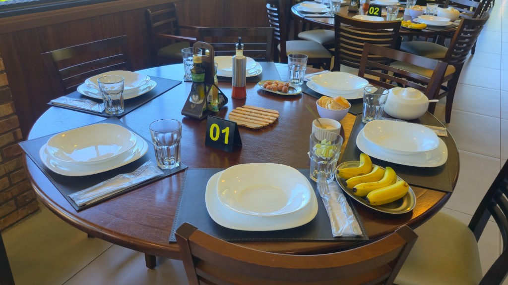 Mesa redonda de madeira escura posta para seis pessoas com pratos brancos, copos, talheres embalados, azeite, pães, molhos, bananas e salgadinhos, em restaurante com outras mesas ao fundo