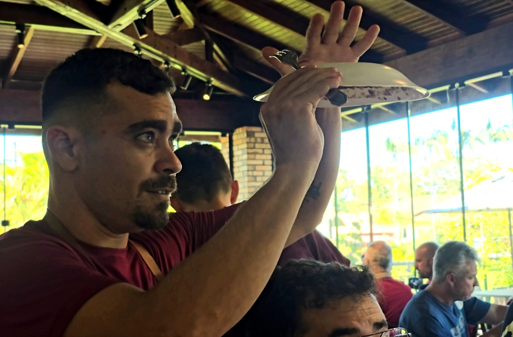 Homem de barba e camiseta vinho segura um prato branco com uma colher, olhando para cima. Ao fundo, outras pessoas e uma janela com vegetação externa