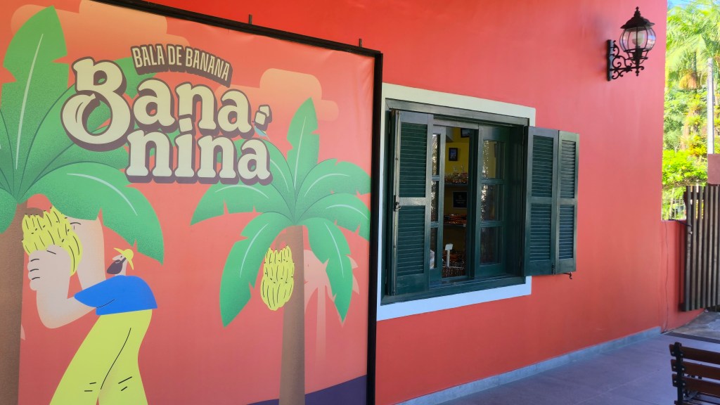 Fachada de uma casa com parede laranja avermelhada e um banner da Bala de Banana Bananina. O banner mostra um homem colhendo bananas de uma bananeira, com o nome do produto em letras grandes. À direita, uma janela de madeira verde com venezianas abertas revela o interior da loja. Acima da janela, um lampião preto ornamentado. À direita da casa, vegetação e um portão de madeira vertical. No canto inferior direito, parte de um banco de madeira.