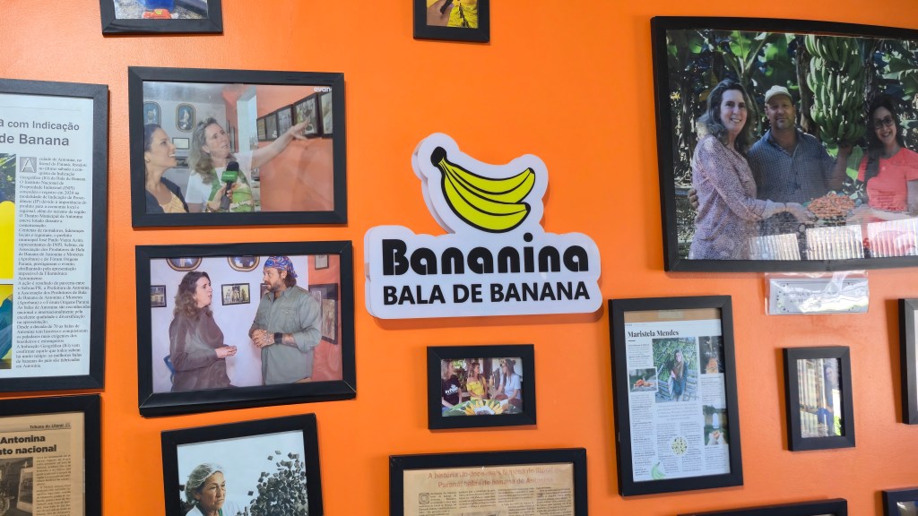 Parede laranja com vários quadros pretos emoldurando fotos e recortes de jornais, e um letreiro branco com bananas amarelas e o texto Bananina Bala de Banana