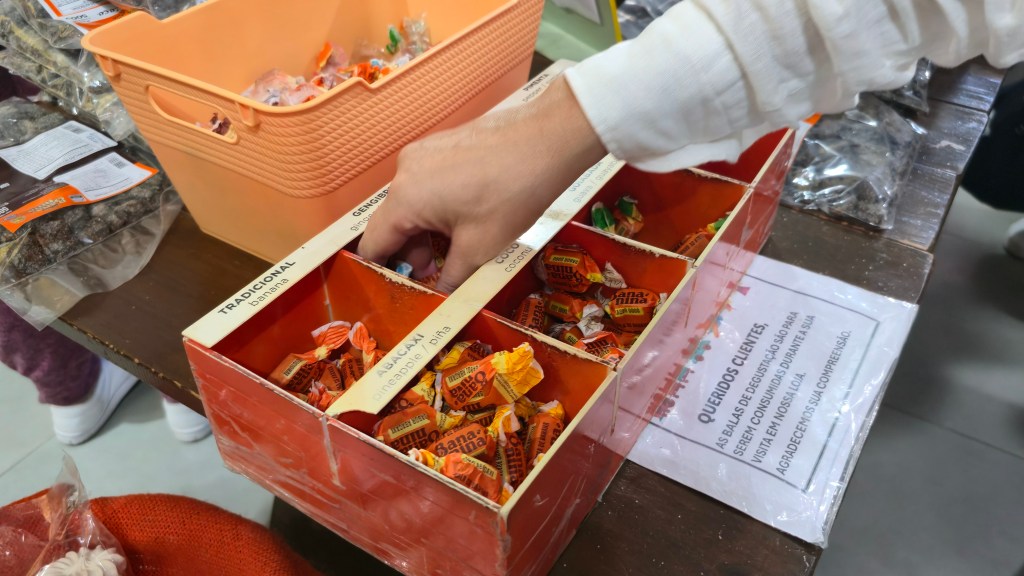 Mão de uma pessoa pegando uma bala de um expositor de madeira com divisórias, contendo balas de banana, abacaxi, coco e gengibre. Ao lado, um aviso para clientes sobre balas de degustação.