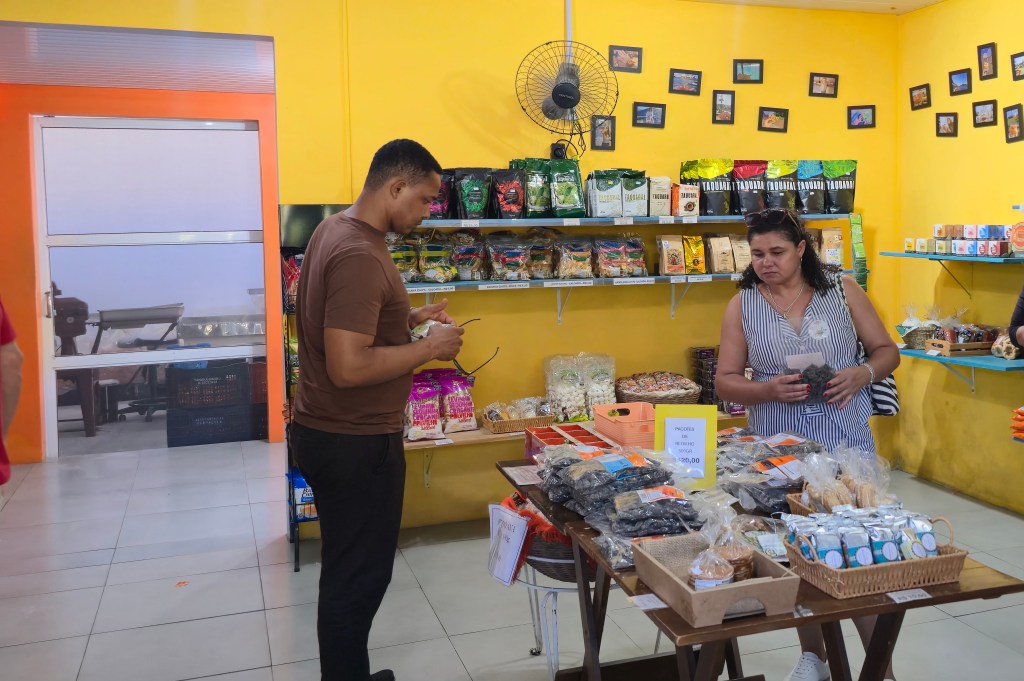 Homem de camiseta marrom e mulher de blusa listrada observam produtos em uma loja com paredes amarelas e prateleiras cheias de pacotes de alimentos