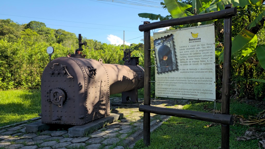 Caldeira a vapor antiga e enferrujada, com um medidor de pressão, sobre um piso de pedras, ao lado de um painel informativo de madeira, em um ambiente verde com árvores e bananeiras