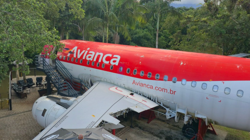 Lateral de um avião Avianca vermelho e branco, com escadas de acesso à fuselagem, em meio a árvores e vegetação densa. A asa esquerda está visível, e ao fundo, um pátio com mesas e cadeiras escuras. O céu azul com nuvens aparece no topo