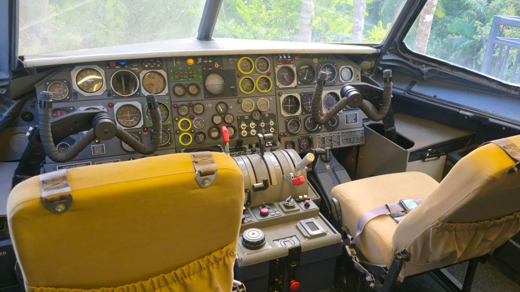 Interior de um cockpit de avião com dois assentos amarelos e bege, painel de controle cinza repleto de botões, alavancas e mostradores, e um manche à esquerda. A janela frontal exibe folhagens verdes