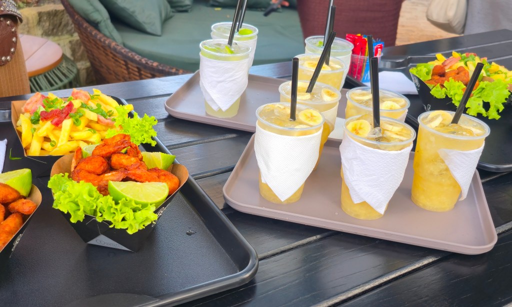 Uma mesa escura com bandejas cheias de petiscos e bebidas. Há porções de batata frita com molho e cebolinha, camarões fritos com alface e limão, e vários copos de suco de banana e limão com canudos pretos. Ao fundo, um sofá de vime com almofadas verdes