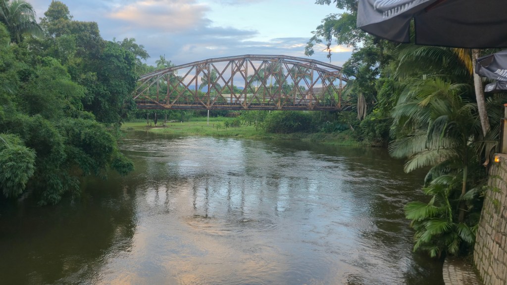 Ponte de ferro enferrujada sobre um rio de águas escuras, cercado por vegetação densa e céu nublado. À direita, um guarda-sol de bar e uma parede de pedras.