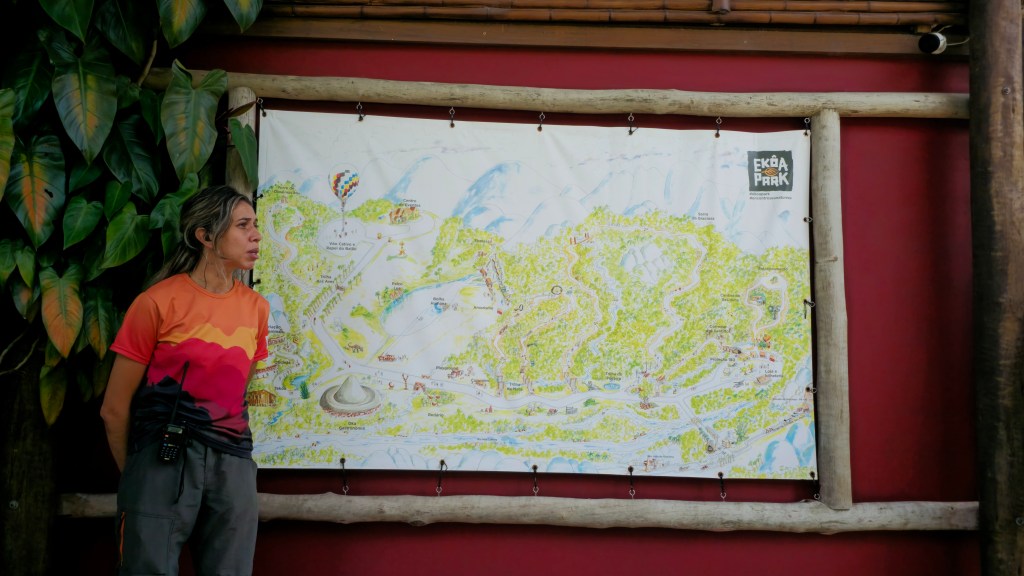 Mulher de cabelo loiro preso, camiseta laranja e roxa, calça cinza, fone de ouvido e rádio na cintura, olhando para um mapa ilustrado do Ekoa Park pendurado em uma parede vermelha. Plantas verdes à esquerda