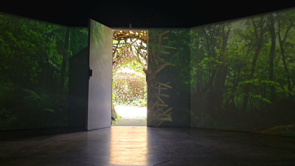 Uma porta branca aberta revela um túnel de galhos e folhagens iluminado, enquanto as paredes laterais exibem projeções de uma floresta densa e verde, criando um ambiente imersivo com o chão escuro refletindo a luz da entrada