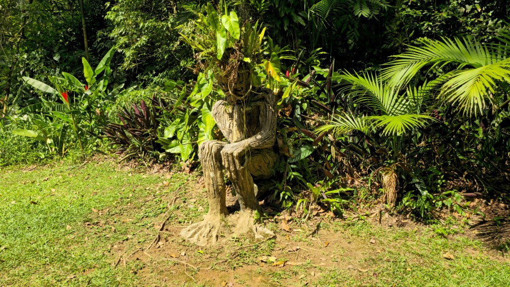 Escultura de tronco de árvore em forma de figura sentada, com folhagens verdes no lugar da cabeça, em meio a uma vegetação exuberante com palmeiras e plantas tropicais, sob luz solar